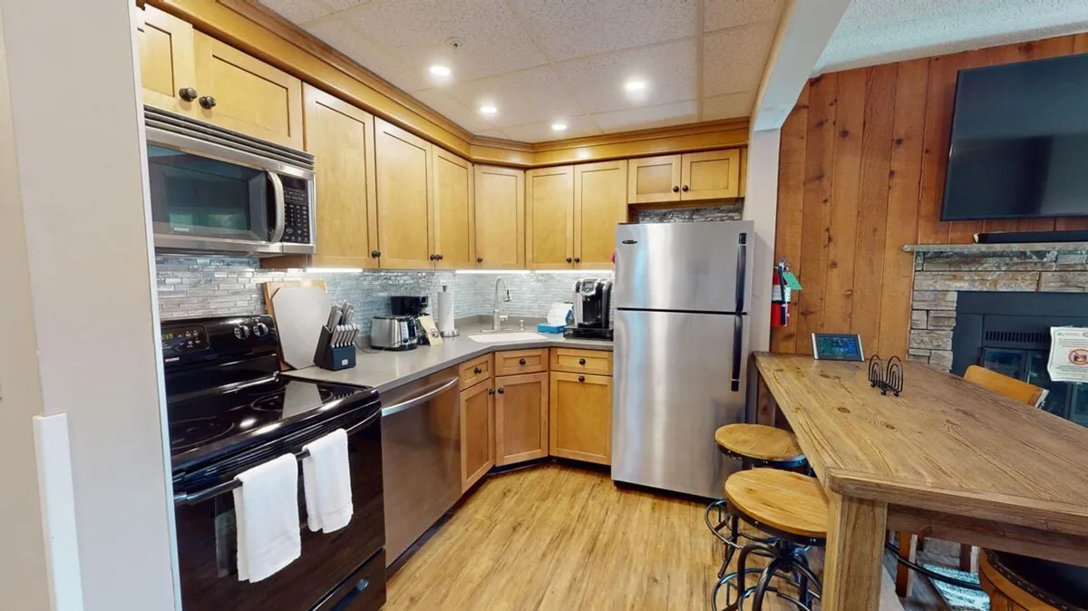 This image offers a view of the kitchen with wood cabinets, modern appliances, bar-style seating, and a nearby fireplace. The setup invites guests to participate in cooking, socializing, or enjoying a quick breakfast at the bar.