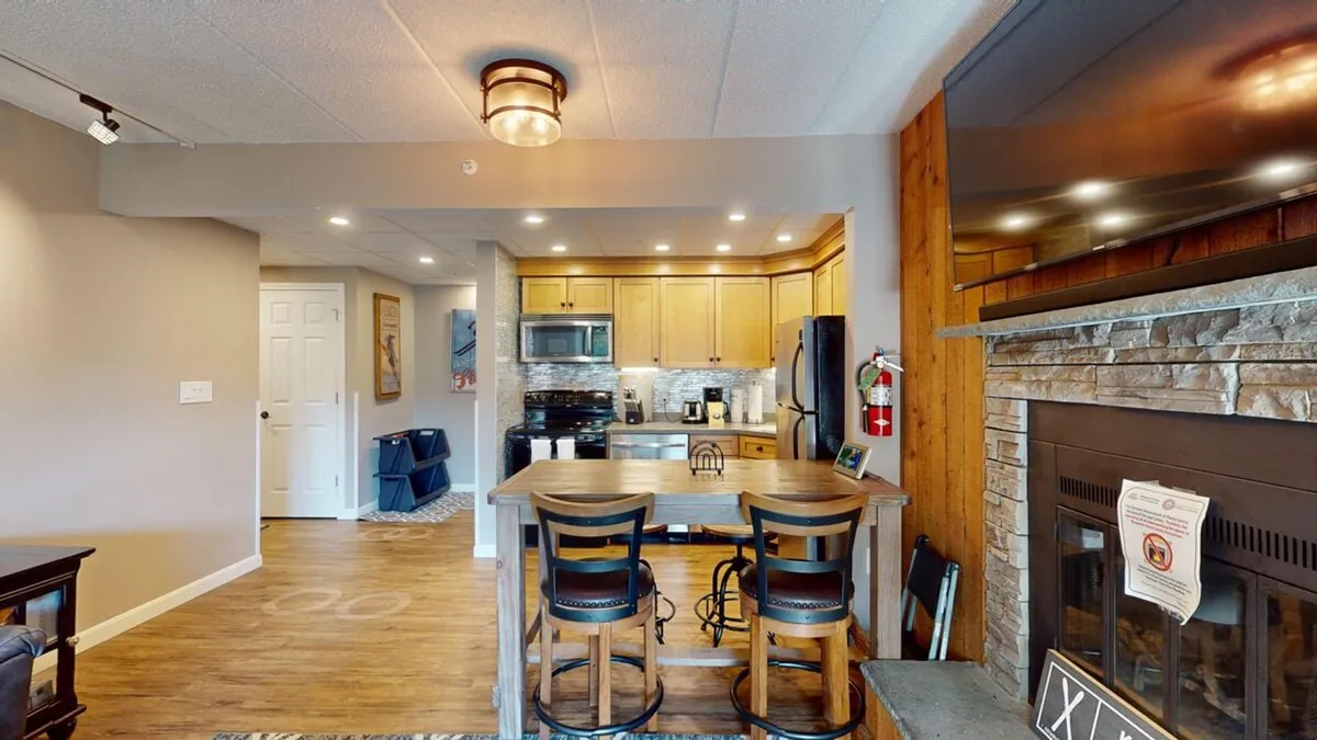 The hero image showcases a warm and inviting open-concept living space featuring a cozy fireplace, a modern kitchen island with stools perfect for casual dining or gatherings, sleek kitchen appliances, and eye-catching mountain-inspired artwork on the wall. The scene embodies relaxation and togetherness, ideal for families or groups seeking comfort after a day on the slopes.