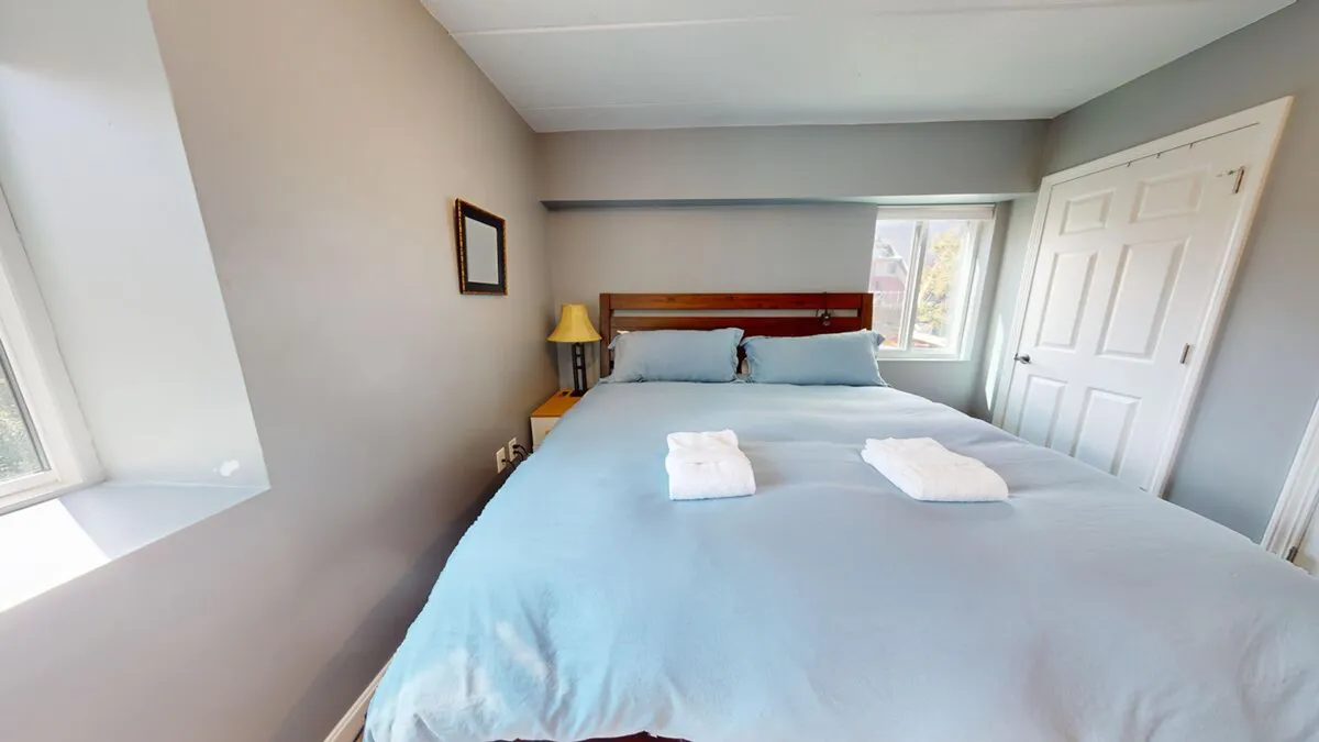 A serene bedroom featuring a comfortable bed with light blue bedding, a nightstand with a lamp, a window letting in abundant natural light, and a framed mirror. The space looks peaceful and inviting, ideal for unwinding at the end of a busy day.