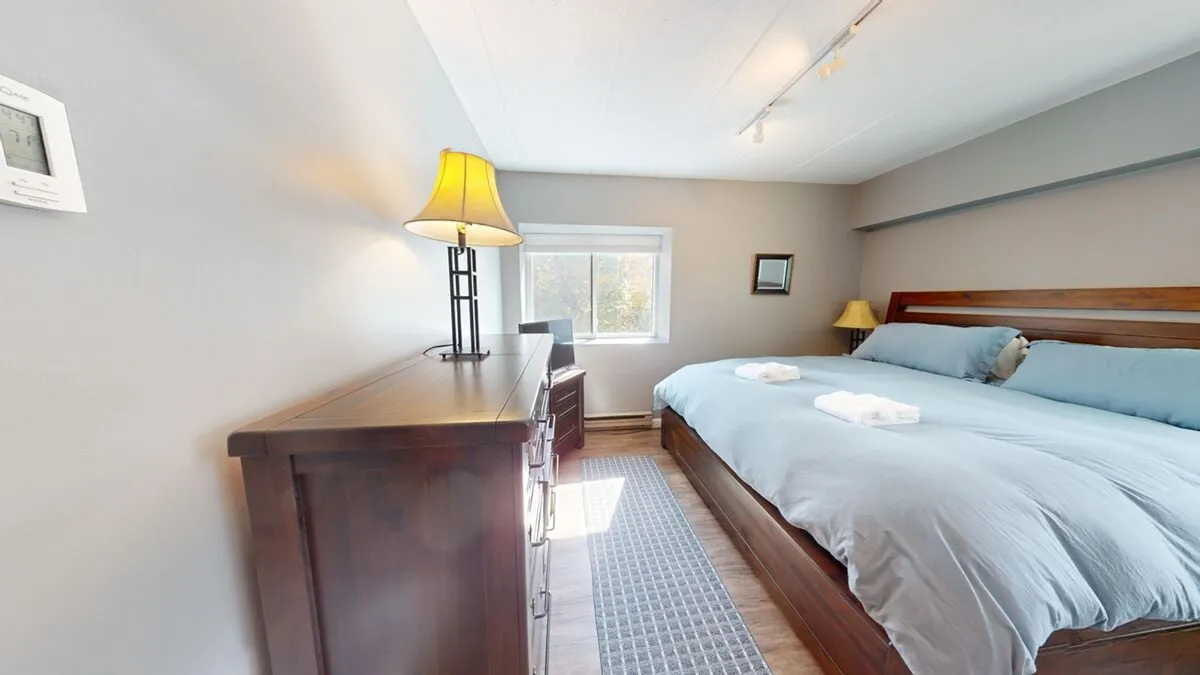 A serene bedroom with blue linens on the bed, a wooden dresser, a lamp, and a window letting in natural light. The room conveys peace and comfort, emphasizing restful moments and enjoying views beyond the window.