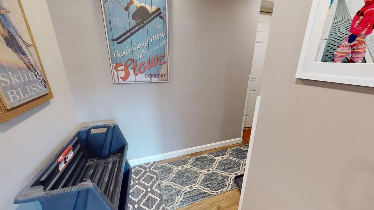 A view of the condo entrance area decorated with a skiing poster, a rug, and a storage bin for equipment. The scene suggests guests coming in or heading out for outdoor activities, providing a strong connection to the winter sports narrative.
