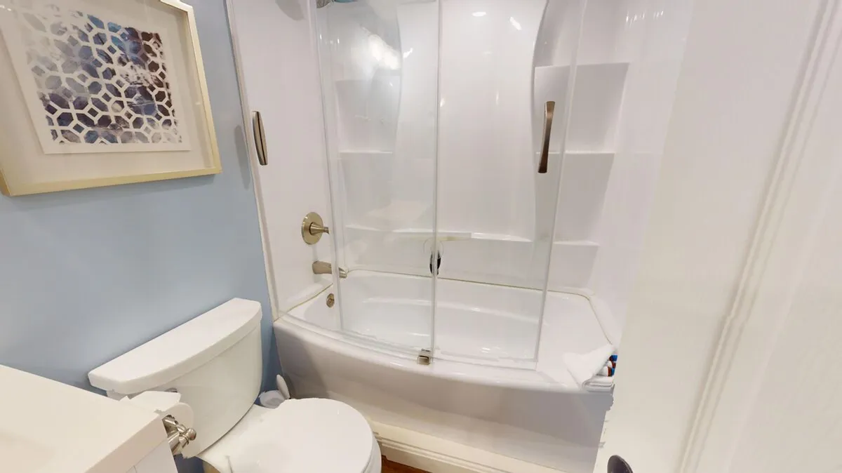 A calming bathroom with a bathtub and glass shower doors, complemented by a toilet, framed artwork, and soothing light blue walls. The space feels refreshing and well-appointed, inviting for both morning routines and evening relaxation.