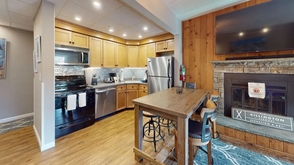 This image showcases the core of the open-plan living space, featuring the kitchen cabinetry, modern stainless steel appliances, a large dining table, and a cozy fireplace. The setup encourages both cooking and socializing, highlighting the kitchen as the heart of the home.